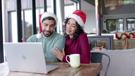 Happy-biracial-couple-wearing-santa-claus-hats-using-laptop-for-video-call-at-home,-in-slow-motion