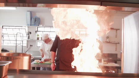 Focused-caucasian-male-chef-frying-food-in-frying-pan-with-bursting-fire-in-kitchen,-slow-motion