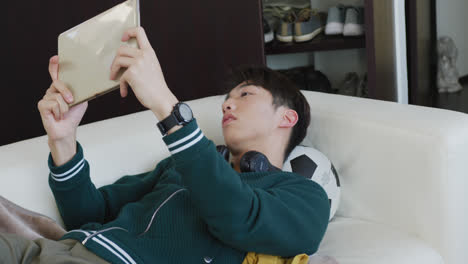 Asiatischer-Junge-Spielt-Spiele-Auf-Einem-Digitalen-Tablet,-Das-Zu-Hause-Auf-Der-Couch-Liegt