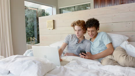 Happy-diverse-gay-male-couple-lying-on-bed-and-using-smartphone-and-laptop-at-home,-slow-motion