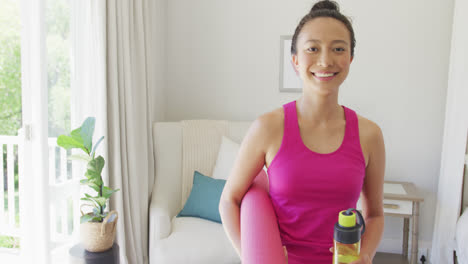 Portrait-of-happy-asian-woman-holding-yoga-mat-and-water-bottle-in-bedroom,-in-slow-motion