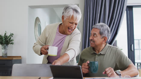 Happy-senior-biracial-couple-talking-with-coffee-and-using-laptop