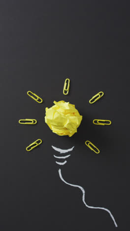 Vertical-video-of-yellow-paper-ball-and-paperclips-forming-lightbulb-on-black-background
