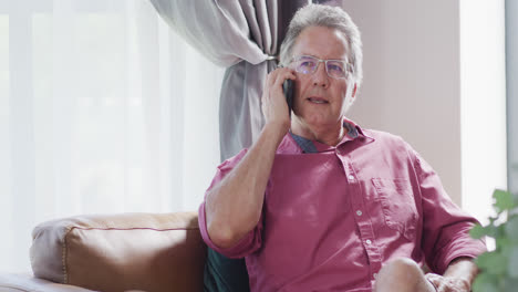 Happy-senior-caucasian-man-sitting-on-sofa-and-talking-on-smartphone,-slow-motion