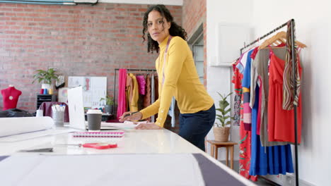 Portrait-of-happy-biracial-female-fashion-designer-standing-at-table-working-in-studio,-slow-motion