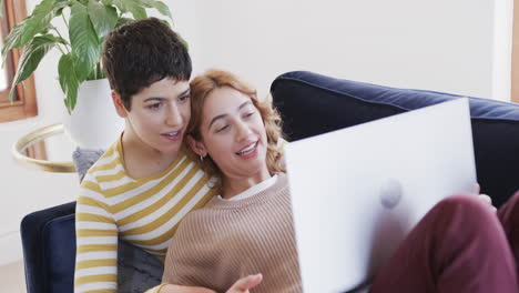 Happy-caucasian-lesbian-couple-lying-on-sofa,-using-laptop-and-smiling-in-sunny-house