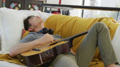 Asian-male-teenager-with-headphones-playing-guitar-and-lying-in-living-room
