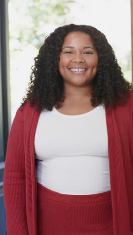 Vertical-video-of-happy-african-american-woman-with-curly-hair-standing-and-smiling-in-sunny-house