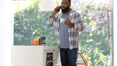 African-american-man-drinking-coffee-and-talking-on-smartphone-in-sunny-kitchen,-slow-motion
