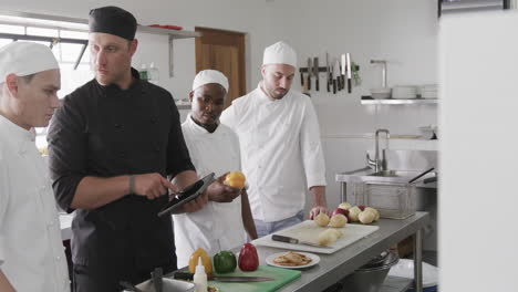 Chef-Masculino-Diverso-Instruyendo-A-Un-Grupo-De-Chefs-Masculinos-En-Prácticas-Usando-Tableta-En-La-Cocina,-Cámara-Lenta