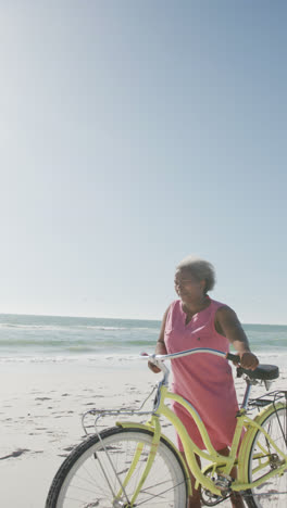 Vertical-video-of-happy-senior-african-american-woman-wlaking-with-bike-at-beach,-in-slow-motion