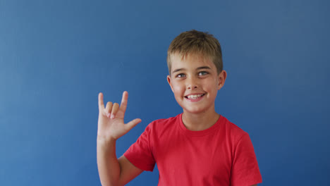Niño-Caucásico-Feliz-Usando-Lenguaje-De-Señas-Y-Sonriendo-Sobre-Fondo-Azul,-Cámara-Lenta