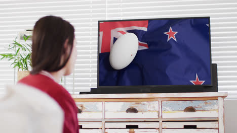 Caucasian-woman-watching-tv-with-rugby-ball-on-flag-of-new-zeland-on-screen