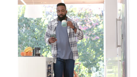 African-american-man-drinking-coffee-and-using-smartphone-in-sunny-kitchen,-slow-motion