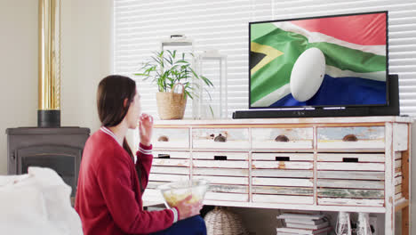 Caucasian-woman-watching-tv-with-rugby-ball-on-flag-of-south-africa-on-screen