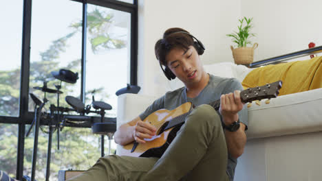 Asian-boy-wearing-headphones-playing-guitar-in-living-room-at-home