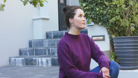Happy-caucasian-woman-in-purple-sweater-sitting-in-front-of-house-in-garden