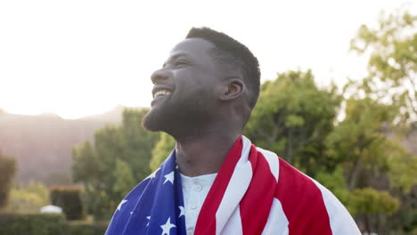 Glücklicher-Afroamerikanischer-Mann,-Der-Mit-Flagge-Auf-Dem-Rücken-Steht-Und-Im-Sonnigen-Garten-Lächelt