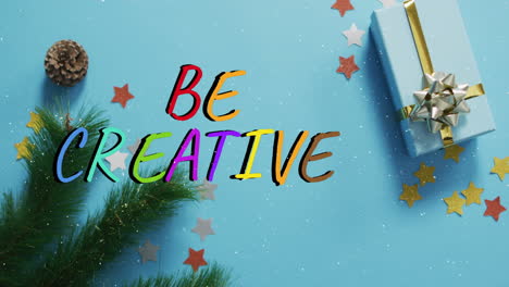 Animation-of-snow-falling-over-be-creative-text-banner-against-gifts-and-decorations-on-blue-surface