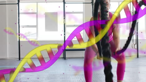 Animation-of-dna-strands-over-happy-caucasian-woman-cross-training-with-battle-ropes-at-gym