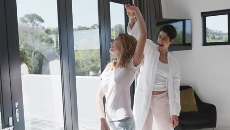 Happy-caucasian-lesbian-couple-dancing-by-window-and-embracing-in-sunny-house