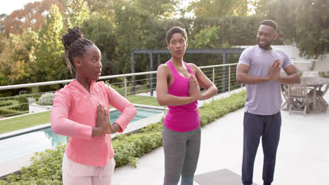 Felices-Padres-Afroamericanos-E-Hija-Practicando-Yoga-En-Un-Jardín-Soleado,-En-Cámara-Lenta