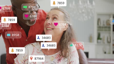 Animation-of-social-media-icons-and-numbers-over-senior-diverse-couple-at-home
