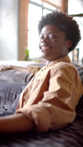 Vertical-video-of-portrait-of-happy-african-american-man-sitting-on-sofa-at-home,-slow-motion