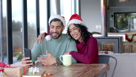 Happy-biracial-couple-wearing-santa-claus-hats-using-smartphone-for-video-call-at-home,-slow-motion