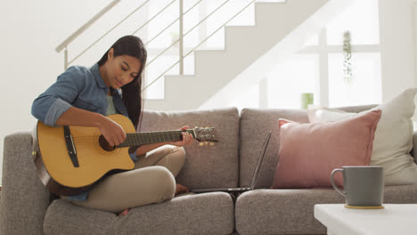 Video-of-happy-biracial-woman-sitting-on-sofa-and-playing-guitar