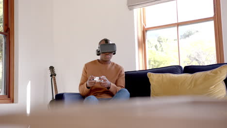 African-american-man-sitting-on-sofa-using-vr-headset-and-playing-video-games-at-home,-slow-motion
