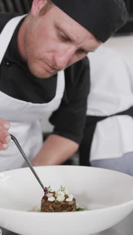 Focused-caucasian-male-chef-decorating-prepared-meal-on-plate-in-kitchen,-slow-motion,-vertical