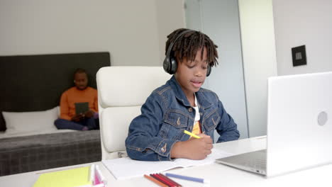 Niño-Afroamericano-Usando-Una-Computadora-Portátil-Para-Una-Lección-En-Línea-Con-Su-Padre-En-Segundo-Plano-En-Casa,-Cámara-Lenta