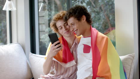Diverse-gay-male-couple-with-rainbow-lgbt-flag,-using-smartphone-on-sofa-at-home,-slow-motion