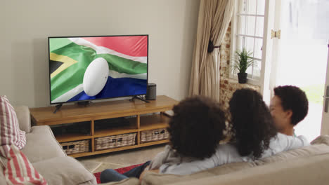 Gemischtrassige-Familie-Vor-Dem-Fernseher-Mit-Rugbyball-Auf-Der-Flagge-Südafrikas-Auf-Dem-Bildschirm