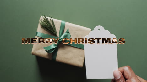 Frohe-Weihnachten-Text-In-Gold-über-Geschenk-Mit-Leeren-Geschenkanhänger-Auf-Grünem-Hintergrund-Mit-Bokeh-Lichtern