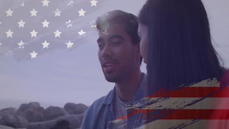 Animation-of-usa-flag-effect-against-biracial-couple-talking-to-each-other-at-the-beach