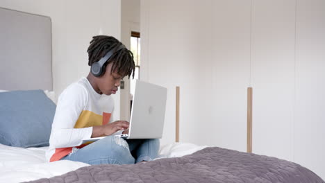 African-american-boy-wearing-headphones-sitting-on-bed-using-laptop-at-home,-slow-motion,-copy-space