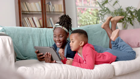 Happy-african-american-sister-an-brother-lying-on-sofa-and-using-tablet-at-home,-slow-motion