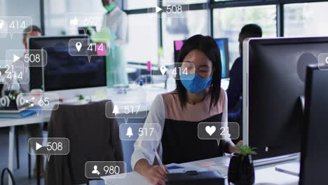 Animation-of-social-media-icons-over-asian-woman-wearing-face-mask-taking-notes-at-office