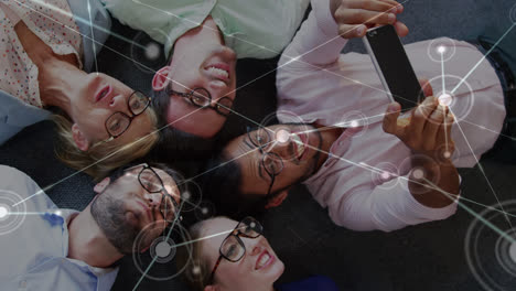 Animation-of-dots-connecting-with-lines-over-man-taking-selfie-with-coworkers-while-lying-on-floor