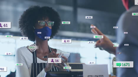 Animation-Von-Social-Media-Symbolen-Und-Daten-über-Einer-Afroamerikanischen-Verkäuferin-Mit-Gesichtsmaske