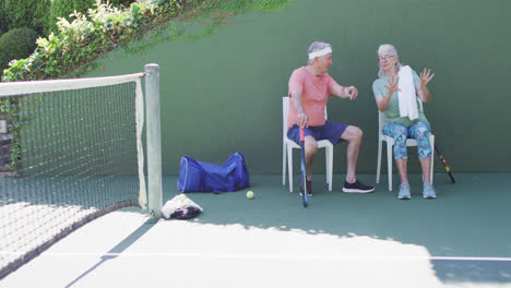Happy-senior-caucasian-couple-taking-a-break,-sitting-talking-at-outdoor-tennis-court,-slow-motion