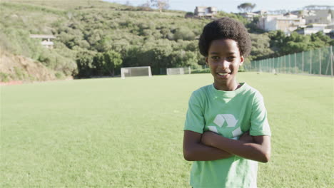 Un-Niño-Birracial-Se-Encuentra-Con-Confianza-En-Un-Campo-De-Fútbol,-Vistiendo-Una-Camiseta-Verde-De-Reciclaje,-Con-Espacio-Para-Copiar