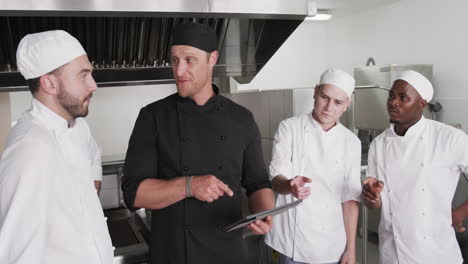 Diverse-male-chef-instructing-group-of-trainee-male-chefs-using-tablet-in-kitchen,-slow-motion