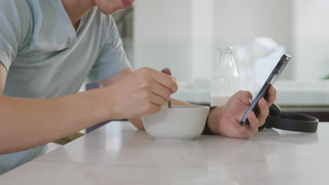 Asiatischer-Männlicher-Teenager,-Der-Mit-Smartphone-Am-Tisch-Sitzt-Und-Frühstückt