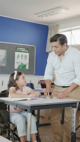 Vertical-video-of-happy-diverse-male-teacher-and-schoolgirl-in-wheelchair-writing-in-slow-motion