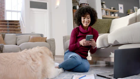 Glückliche-Biracial-Frau-Mit-Golden-Retriever-Hund-Mit-Laptop-Und-Smartphone-Zu-Hause,-Zeitlupe