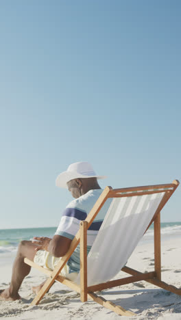 Vertical-video-of-senior-african-american-man-using-laptop-on-deckchair-at-beach,-in-slow-motion