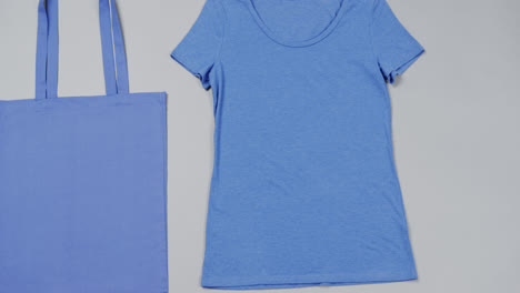 Primer-Plano-De-Bolsa-Azul-Y-Camiseta-Sobre-Fondo-Gris,-Con-Espacio-Para-Copiar,-Cámara-Lenta